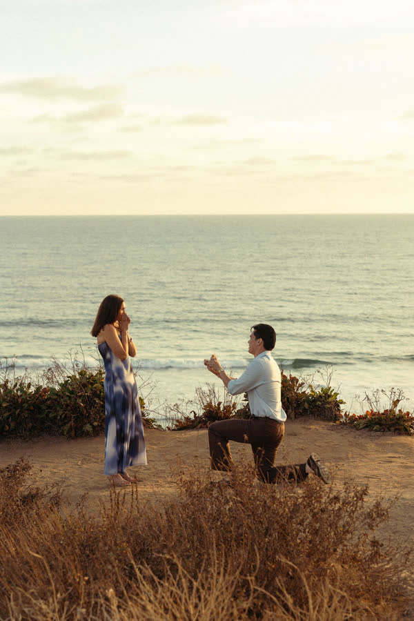 Dylan on one knee proposing to Jessica on a clifftop above the ocean