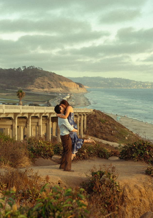 Dylan lifting Jessica into a kiss on the cliff path, the coastline and bridge behind