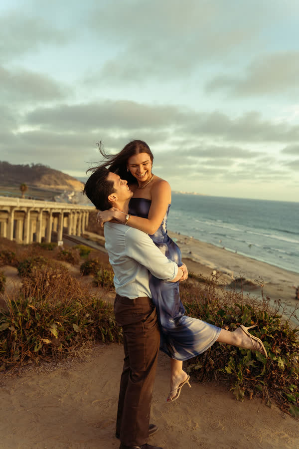 Jessica laughing as Dylan lifts her, wind in her hair, the beach below